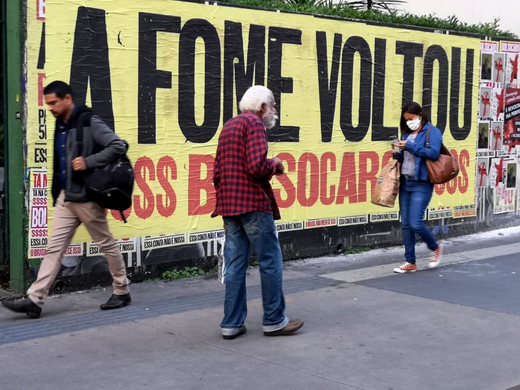 Pessoas passam diante de um outdoor que diz que a fome voltou ao brasil - foto: A Fome Voltou. Lambe lambe em muro na Avenida Paulista, altura da rua Haddock Lobo. S&atilde;o Paulo, SP. 16 de abril de 2021. Foto: Roberto Parizotti.Ag&ecirc;ncia Fotos Publicas